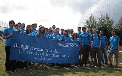 A large group of UoS Roadshow exhibitors on the grass outside the Winchester Science Centre, wearing a branded roadshow blue t-shirt and holding a blue roadshow banner.