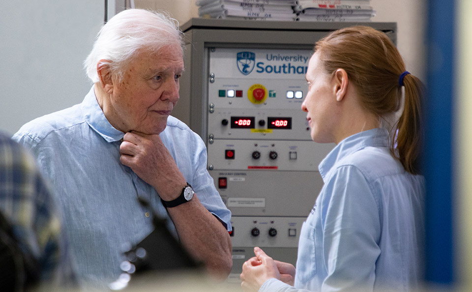 Woman scientist in blue shirt speaking to TV presenter Sir David Attenborough in a lab.