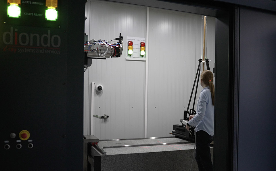 Scientist in a large room sized x-ray scanner standing near a metal scanning arm.