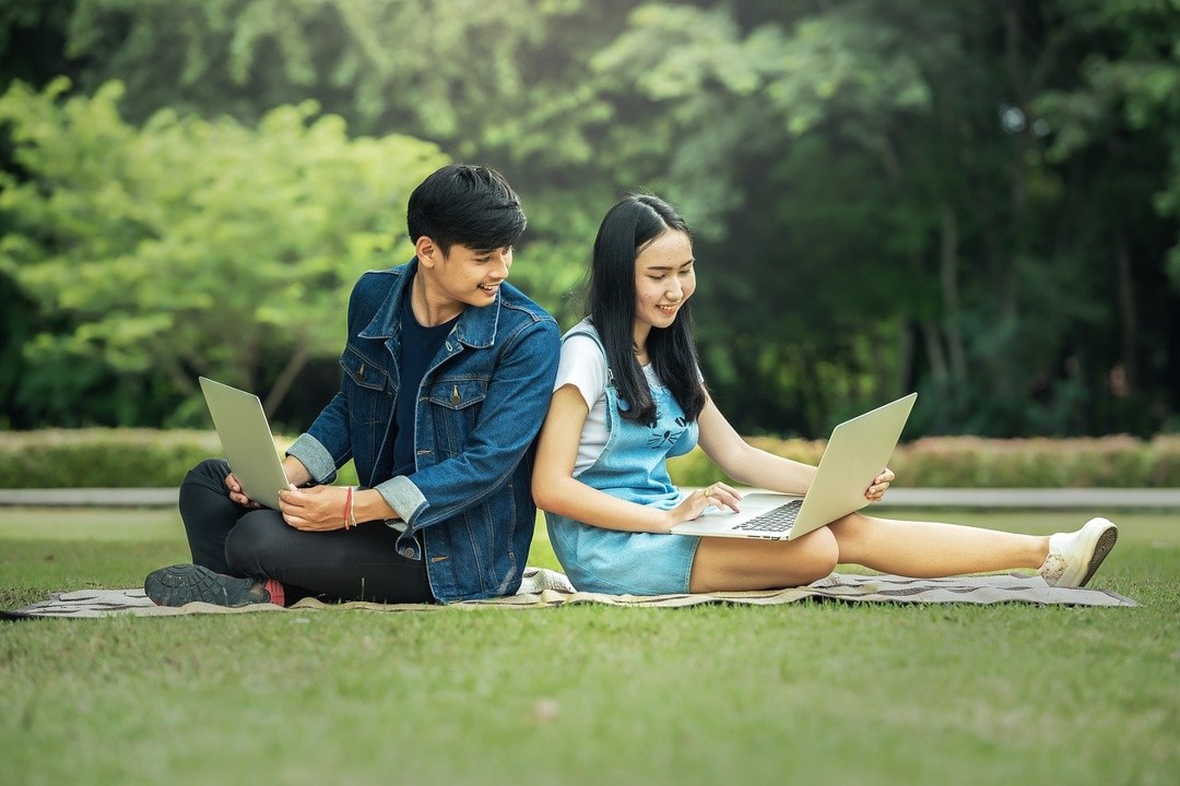 A male and a female student sitting on the grass with laptops.