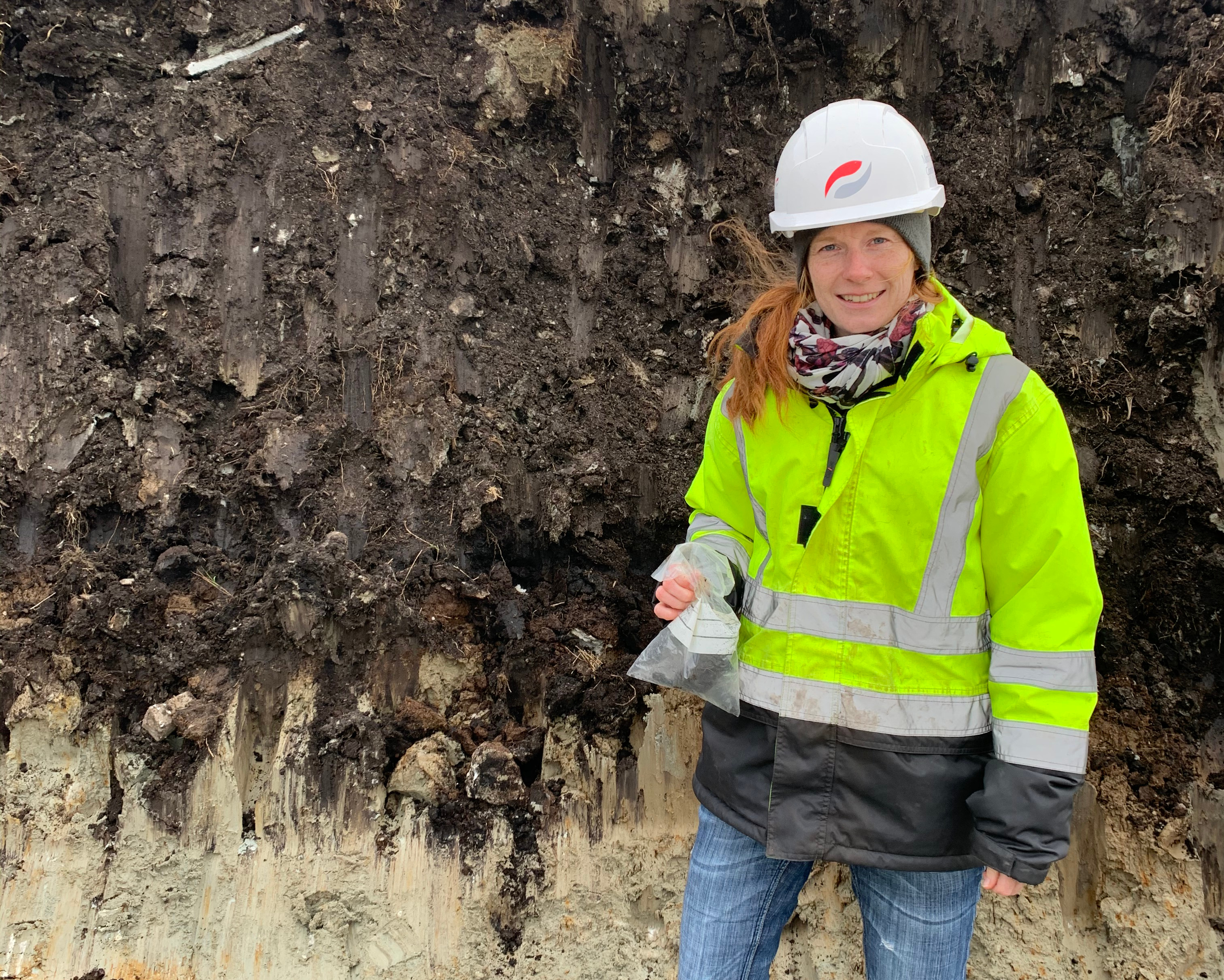 Person in high-visibility jacket and hard hat standing by an exposed soil profile.