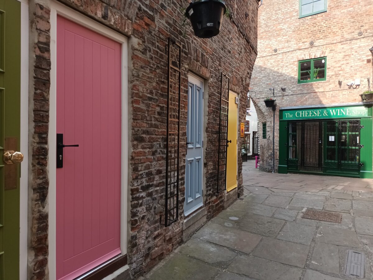 A narrow cobbled alleyway is lined with colourful doors—pink, blue, yellow, and green—set into old brick buildings. At the end of the lane is a green‑fronted shop called “The Cheese & Wine Shop.”