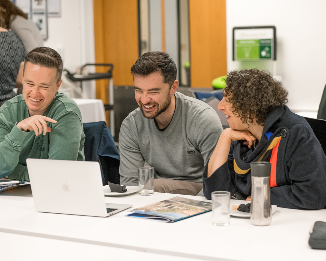 Three people sit at a table looking at a laptop, with notebooks, glasses of water, and other materials spread out in front of them. The setting appears to be a meeting or workshop space with doors, signage, and equipment visible in the background.