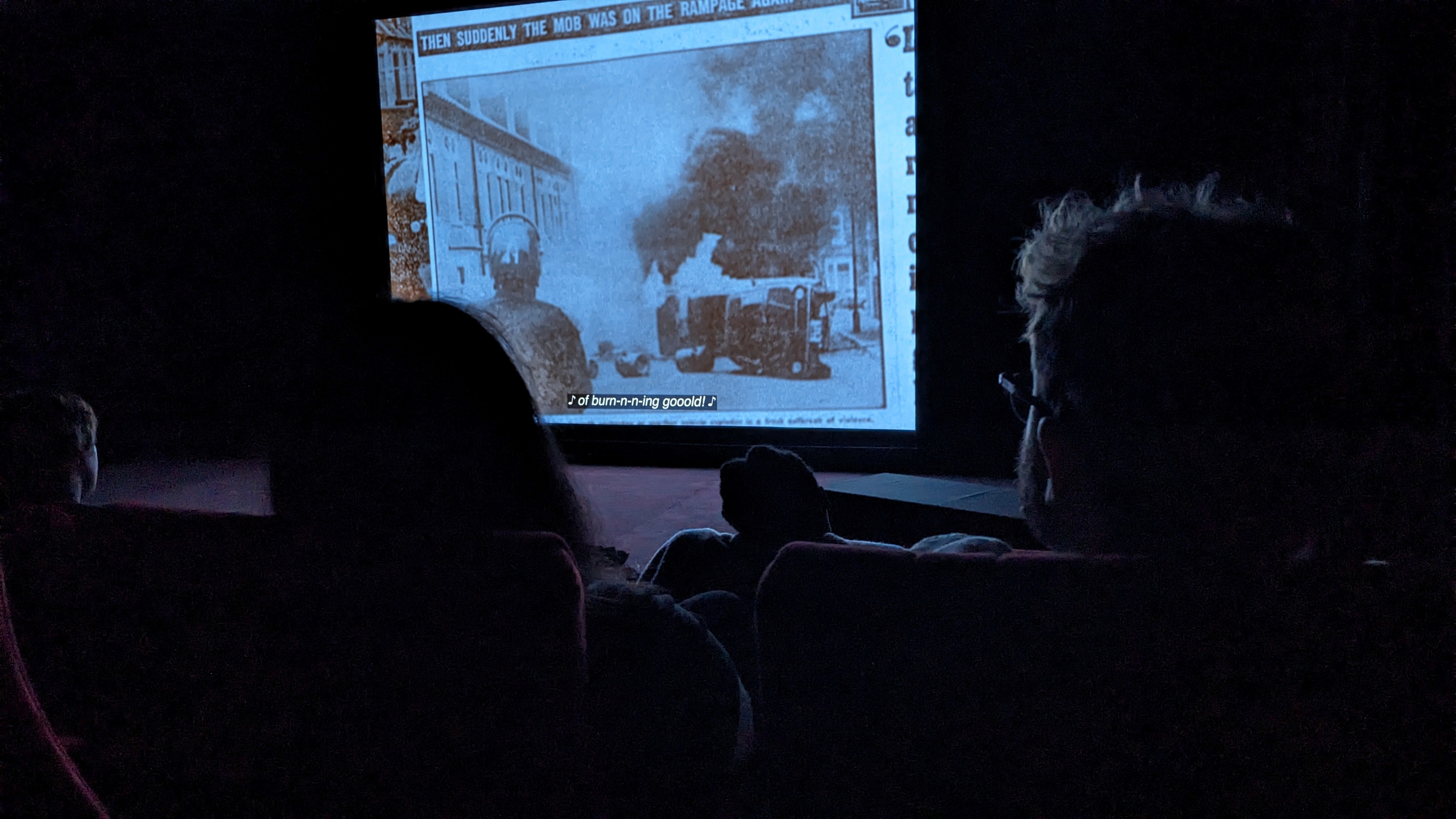 The image shows a dimly lit cinema setting with several people seated in front of a large screen. The screen displays a black-and-white historical photograph of a street scene with smoke rising near vehicles, accompanied by text at the top reading 