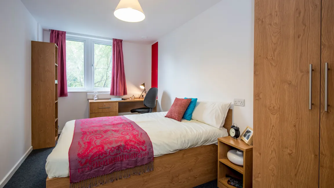 Bedroom with a neatly made double bed and nightstand. A wooden desk and open window are visible in the background.