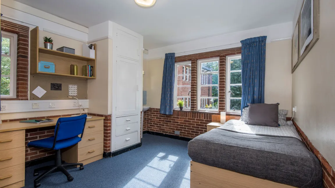 Light from a large window fills a spacious bedroom with desk, chair and neatly made bed.