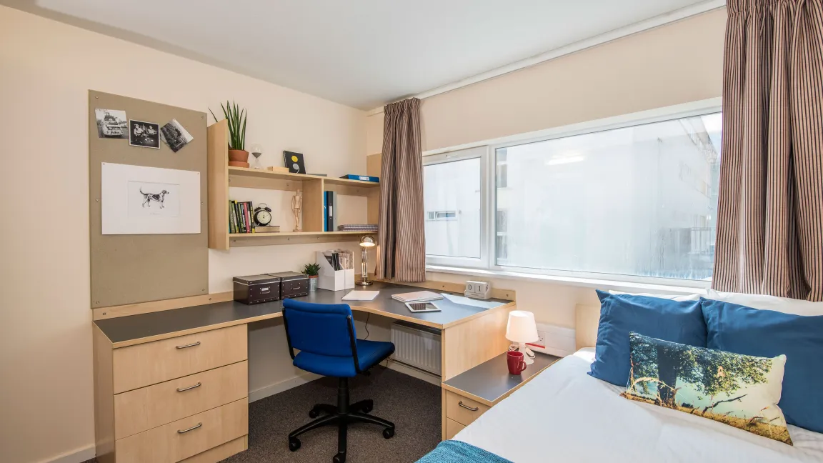 A large window lets light into a bedroom where a neatly made double bed, wash basin and desk can be seen.