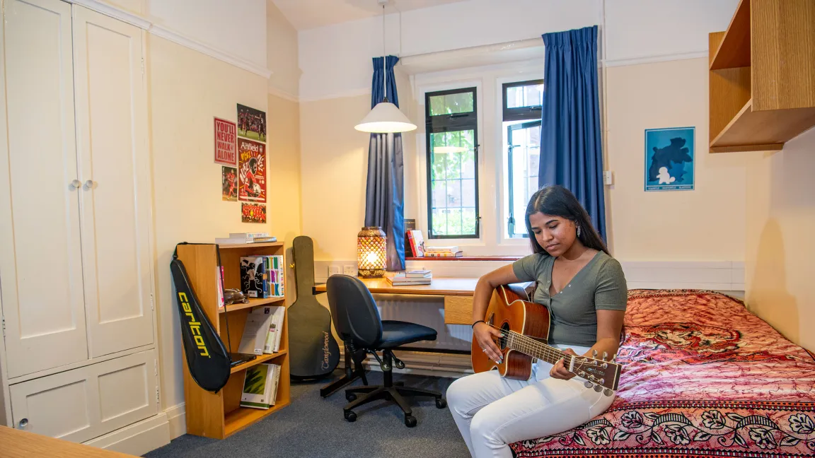 A student plays a guitar while sitting on a bed in a bedroom. In the background, shelves and a desk with books can be seen.