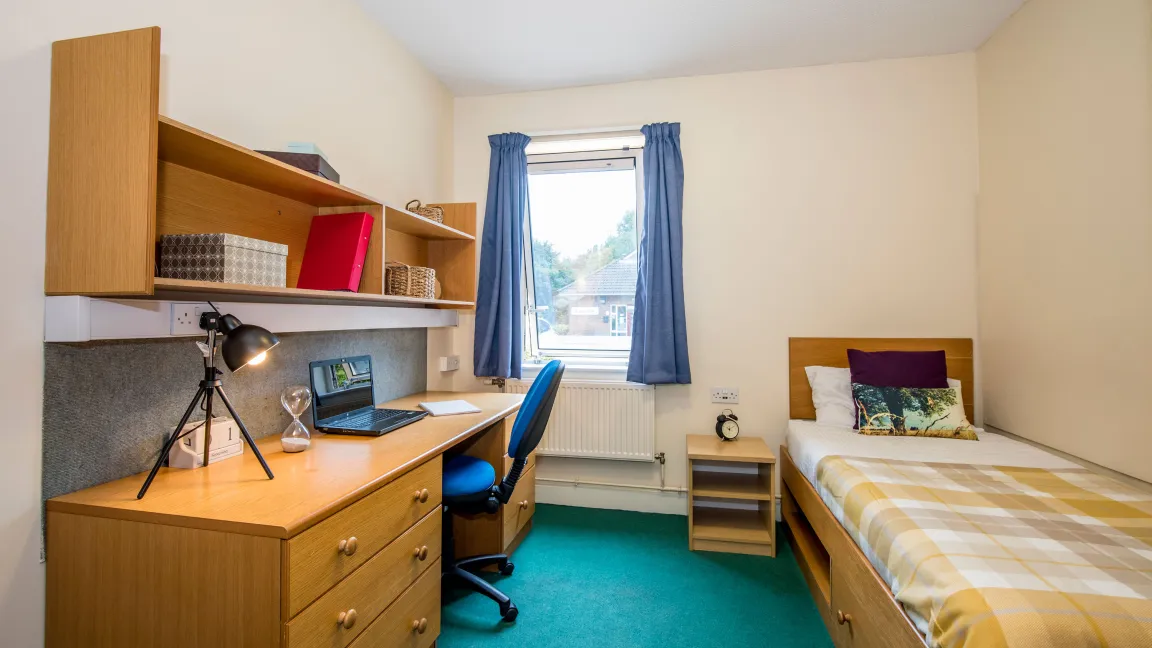 A student bedroom. A section of green carpet separates a single bed with patterned sheets and a large wooden desk. A window on the rear wall lets light in.