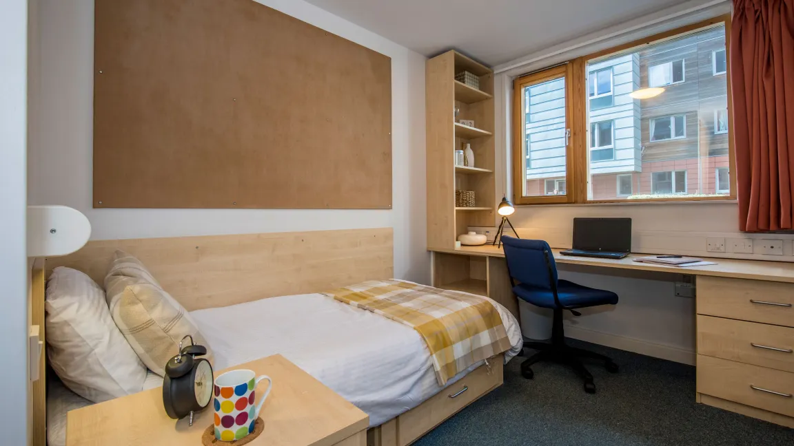 An empty student bedroom containing a neatly made single bed and a large desk with chair, beneath a window.