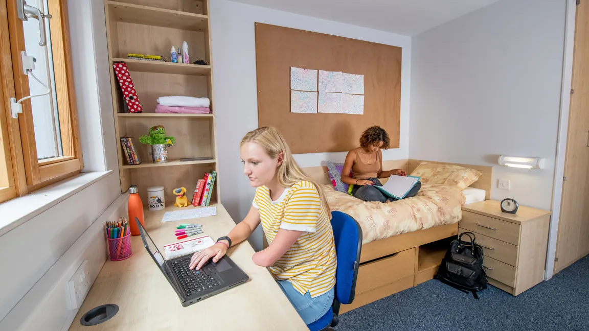 Two students in a bedroom. One sits reading on a single bed while the other works on a laptop, sat at a desk beside a window.
