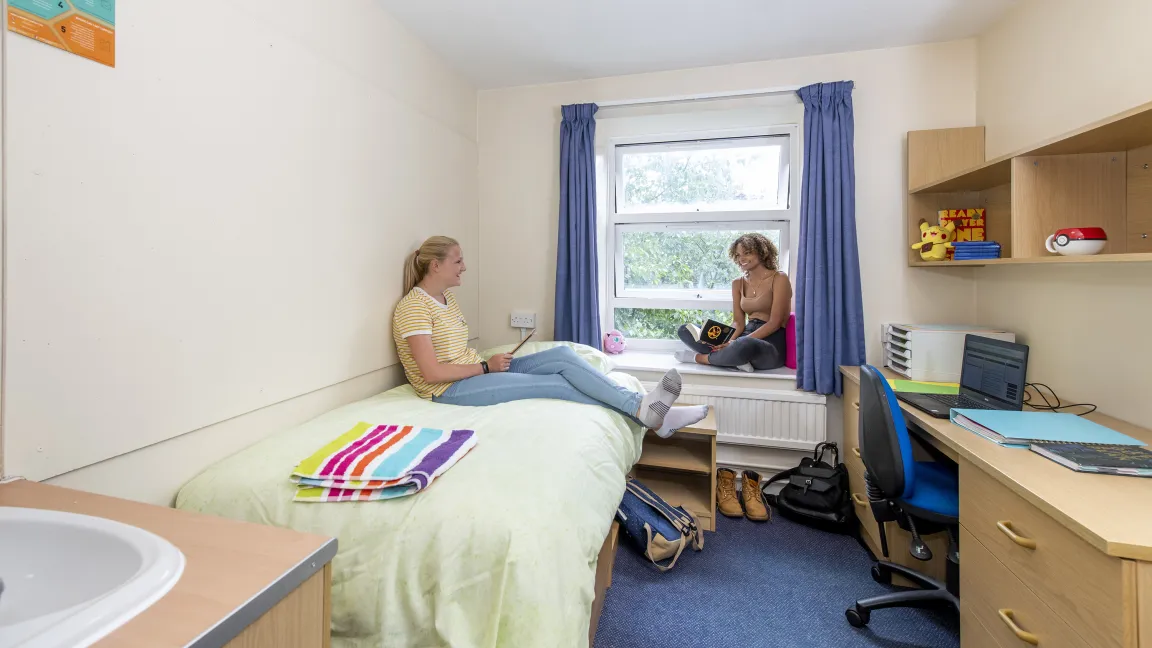Two students chatting in a bedroom. One sits browsing with her tablet on a single bed while the other looks up from their book, sat on the windowsill.