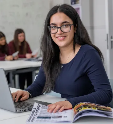 Head and shoulders portrait image of Seerat Chatha, BA Film, TV and Digital Media student smiling at the camera. She has her hands on a laptop and a magazine on the desk in front of her.