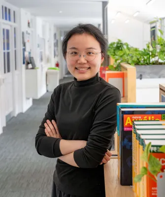 Head and shoulders portrait image of Chang Gao, MA Translation and Professional Communication Skills student smiling and looking at the camera. She is standing with her arms crossed next to a cupboard in a corridor.