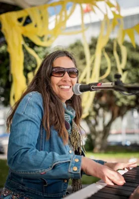Emily Macklow is sitting playing a piano and has a microphone in front of her face.  She is smiling and looking at the camera.