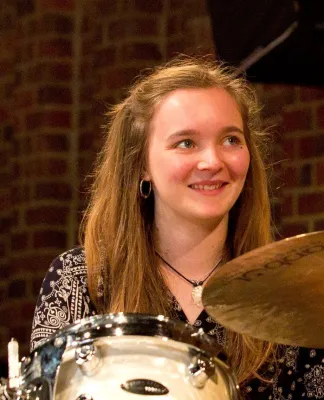  Holly Barnett sitting at her drum kit. She is smiling looking up to the camera.