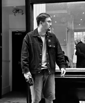 A black and white image of Linhao Li standing outside the glass front of a shop. There are people browsing inside the shop.