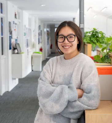 Head and shoulders portrait image of Mengdie Wang, MA Translation and Professional Communication Skills student smiling and looking at the camera. She is leaning on a cupboard with a plant behind her in a corridor.