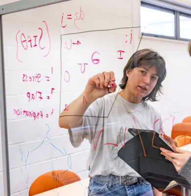 Jodie Adams is writing an equation on a vertical clear board with a red pen.