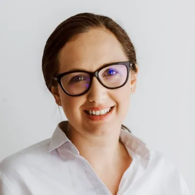 Colour photograph of Sasha Goldstein-Sabbah. Sasha looks directly at the teeth, smiling with teeth showing. She wears her dark hair pulled off her face, and is wearing glasses and a white blouse.