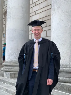 Health Psychology masters student, Nathan is dressed in his graduation robes standing outside a white stonebuilding with columns.