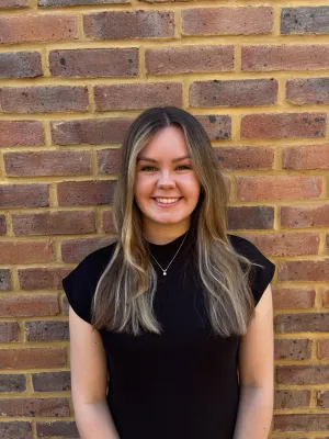 Portrait image of Law student Maddy Nicholl smiling at the camera. There is a red brick wall behind her. 