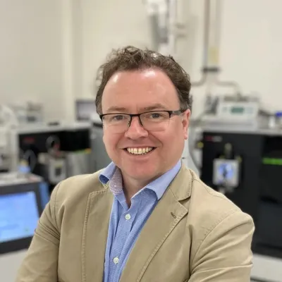 Professor Max Crispin smiling at the camera. He is standing in a lab with a lot of technical equipment in the background.