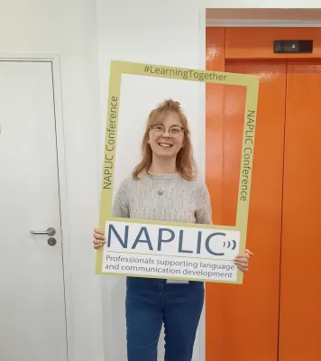 Modern languages and linguistics graduate, Bryony is holding and looking through an open frame. The frame advertises the NAPLIC conference - Professionals supporting language and communication development.