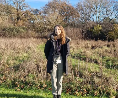 Applied GIS and Remote Sensing master’s student, Jessica, is standing in a field. The grass is long and trees in the background.