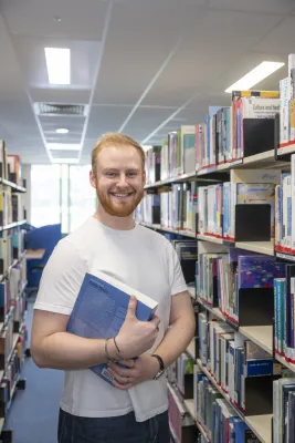 Public Health student, Jack standing between library shelves holding a book. He is smiling at the camera.