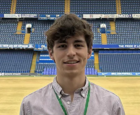 An image of David standing in a large atrium they're using as a night shelter. There a rows of tiered blue seats behind David, who is smiling for the photo.