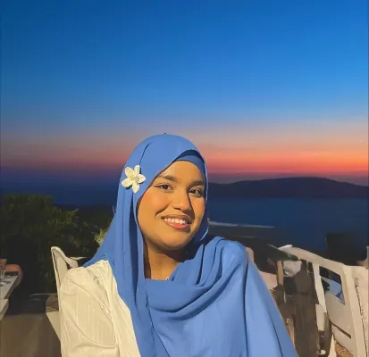 Woman wearing a blue hijab with a flower, smiling at sunset by the sea.