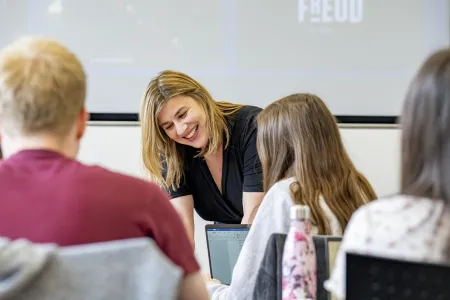 A lecturer smiling while stood at the front of a classroom.