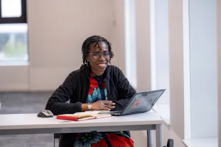 Ogene sat at a desk with a laptop and notebook in front of her. She's looking at the camera and smiling.