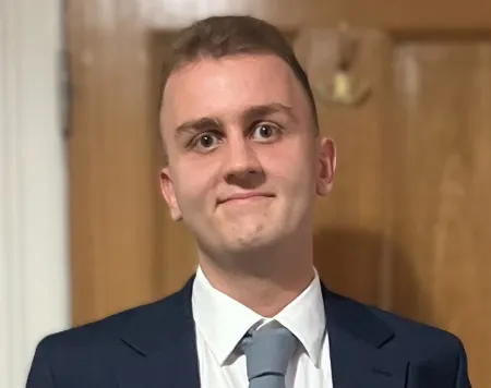 Daniel, a young man with brown hair and eyes and wearing a navy blue blazer and grey tie, smiles at the camera