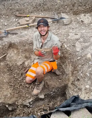 Ben, dressed in reflective orange shorts and wearing gloves, holds a jawbone and smiles proudly at the camera from his dig-site trench