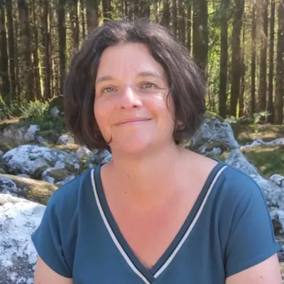 Close-up outdoor portrait of a person with short dark hair wearing a blue top, seated among rocks with tall trees in the background.