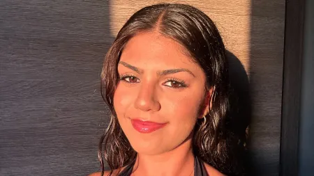 Close-up portrait of a student with dark shoulder-length hair, photographed in warm golden sunlight against a dark textured background.