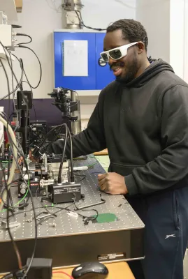Student standing at a bench, adjusts photonics laboratory equipment while wearing protective laser safety glasses.