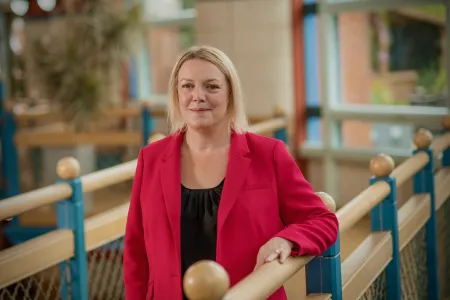 Professor Charlotte Summers, Dean of the Faculty of Medicine. She is wearing a bright red blazer over a block top, standing in a sunny and colourful area surrounded by buildings.