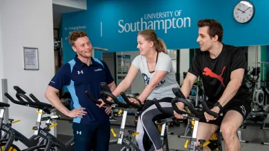 Two students working out on exercise bikes in a gym, assisted by a personal trainer.
