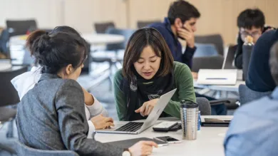 A small group of students sitting around a table look at a laptop screen and discuss their work.