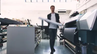 A student walking between several industrial-sized printers and a long work bench lined with guillotine paper cutters
