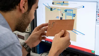 A student compares a prototype PCB to his design on a computer monitor behind it.