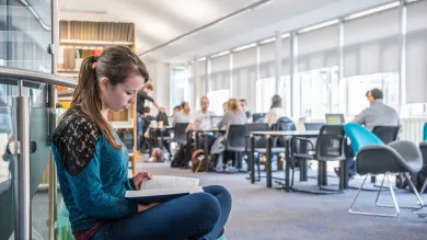 A student sits cross-legged reading a book in a bright, airy library. Behind her are shelves of books and other students working at desks.