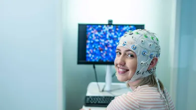 A student sits in front of a computer and turns to smile towards the camera. She is wearing a head cap that is connected to electrodes to monitor brain activity.