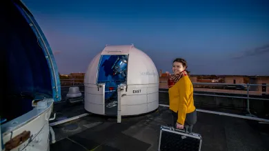 A student stands on the roof of a building at night time. She smiles towards the camera. On the roof are two small dome-shaped observatories containing telescopes.