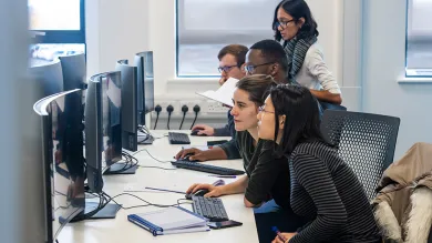 Several students working together across a row of workstations with curved monitors.