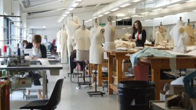 A wide shot of the fashion studio, full of students hard at work. To the right, a row of large workbenches are mixed in with over a dozen mannequins wearing various designs. To the left, a row of sewing machines.