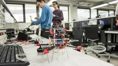 A close-up of a small, 4-rotor drone on a work bench in the laboratory. Two students in the background are testing electronics.